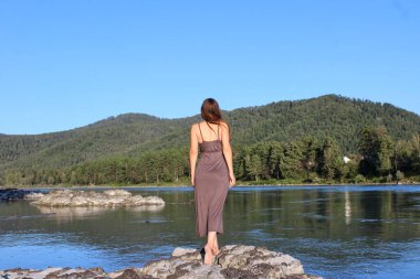 Gri elbiseli, uzun saçlı, güzel vücutlu genç bir kadın güneşli bir yaz gününde nehir kıyısında duruyor. Altai, Sibirya, Rusya. Yeşil tepeler