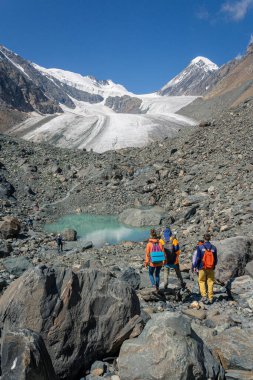 Üç kişilik bir turist grubu, yaz günü Aktru, Altai 'nin yüksek dağları olan küçük bir turkuaz göl ve bir buzula doğru yürüyor. Dikey fotoğraf