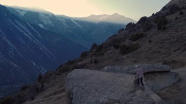 Kadın turist gün batımında dövüş dağlarında dev bir taşın üzerinde yavaşça yürüyor. Hava aracı görüntüsü 4K. Kırgızistan, güzel manzara Ala-Archa Ulusal Parkı bahar alacakaranlığı