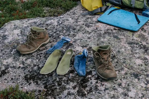 Old brown trekking boots, green insoles and blue socks to dry after day of hiking, laid out on stone. Wet feet while traveling. Travel camping
