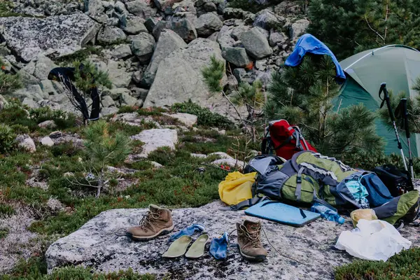 Equipment in the forest: tourist tent, clothes and shoes to dry on tree branches and stones at summer day. Tourist camping on a hike.