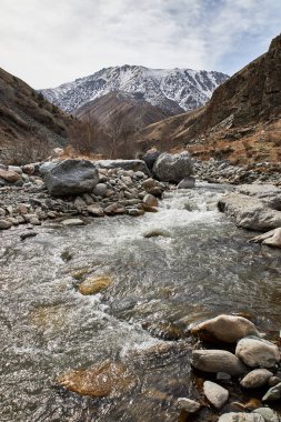 İlkbaharın başlarında doğal manzara. Kar tepeli dağlar, yamaçlar, büyük kayalar ve hızlı akan suyla sığ bir nehrin kayalık kıyısı. Kırgız Cumhuriyeti Kırgızistan dağları