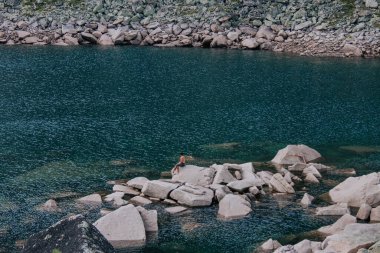 Man sits on large stone protruding from water near shore of emerald-colored mountain lake. Natural landscape, summer hiking day. Hike at kuturchinskoye belogorye Krasnoyarsk region. Tourism in Siberia clipart