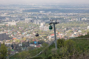 Baharda şehrin arka planına karşı iki kamaralı kablolu bir araba. Almaty, Kazakistan 'daki Kok Tobe tepesine hava nakliyesi. Turistik bir yer, şehir simgesi. Halat destek sütunu