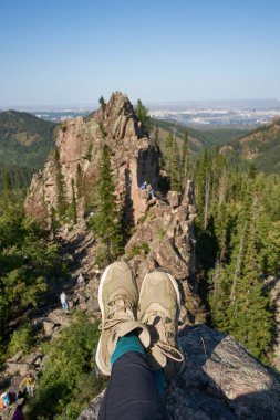 Uçurumun tepesine ulaş. Şehrin yakınlarındaki volkanik kayaların ve yeşil ormanın arka planında yürüyüş botları giymiş bir adamın bacakları. Sigortasız olarak kayanın kenarında oturmanın tehlikesi. Şaşırtıcı doğa.