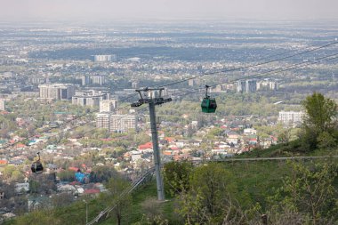 Baharda şehrin arka planına karşı iki kamaralı kablolu bir araba. Almaty, Kazakistan 'daki Kok Tobe tepesine hava nakliyesi. Turistik bir yer, şehir simgesi. Halat destek sütunu