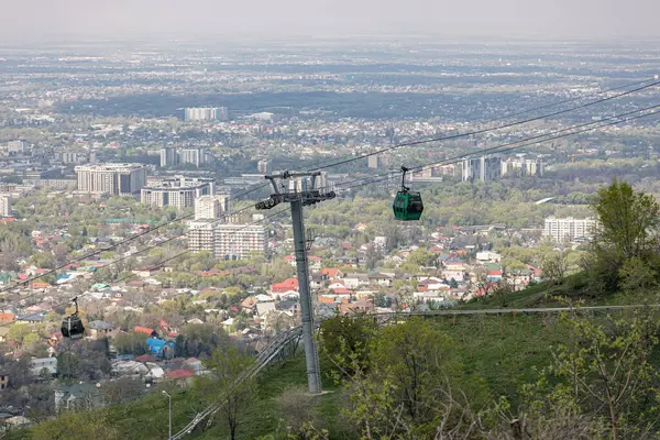 Baharda şehrin arka planına karşı iki kamaralı kablolu bir araba. Almaty, Kazakistan 'daki Kok Tobe tepesine hava nakliyesi. Turistik bir yer, şehir simgesi. Halat destek sütunu