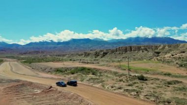 Drone siyah minivan yan görüntüsünü takip ediyor. Araba toprak yoldan sola dönüyor, bulutlu dağlık alana doğru. Yaz günü, doğal manzara, kırsal alan. 4k hava aracı, Kırgızistan seyahat ediyor.