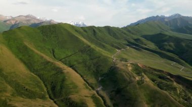Dağ geçidinden geçen dolambaçlı bir yol, kırsal kesimde zor bir yol. Kırgızistan 'ın doğası: öndeki yeşil tepeler ve uzaktaki karlı tepeler. Yaz dağları doğal hava manzarası