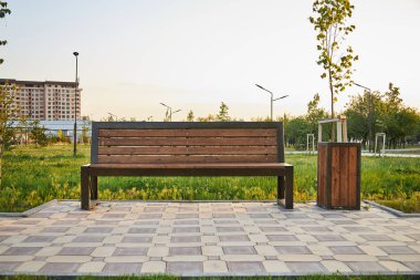 Tranquil Park 'taki metal bir çöp kutusuyla eşleştirilmiş ahşap bank. Güneş batarken ılık bir ışık kareli kaldırımda uzun gölgeler bırakıyor. Çimenler ve genç ağaçlar, huzurlu bir ortam. şehir gelişimi