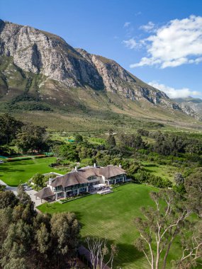 Big house, building, bright green lawn, hotel territory, blue sky. October in Hermanus Klein River Lagoon, Kleinriversvlei, Riversvlei, South Africa. High rocky mountain. Aerial drone photo.  clipart