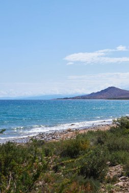 Stone coast of southern shore of big lake Issyk-Kul, summer vacation in Kyrgyzstan. blue sky, emerald colored water of pond, small wave. Kirgiziya travel destination clipart