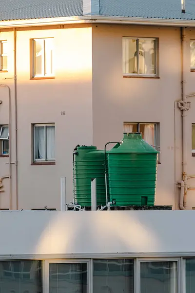 İki büyük, yeşil plastik plastik su tankı çok katlı bir binanın arka planına karşı düz bir çatıda konumlandırılmıştır.