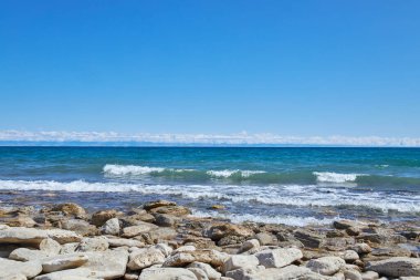 Issyk-Kul Gölü, taş kıyısı. Yaz doğal manzarası, Kırgızistan 'da tatil. Açık mavi gökyüzü, zümrüt renkli gölet suyu, küçük dalga. Kirgiziya seyahat rotası