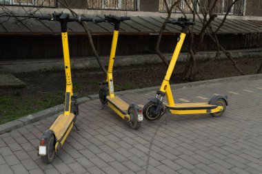 Parke taşlarının üzerinde kiralık üç sarı elektrikli scooter var.