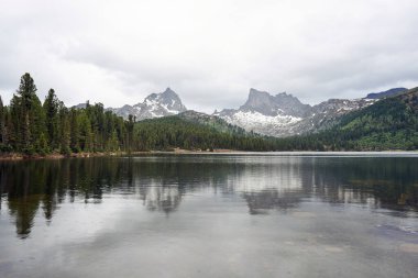 Bahar dağları manzarası. İki dağ zirvesi, kozalaklı orman ve göl, bulutlu gökyüzü.. 