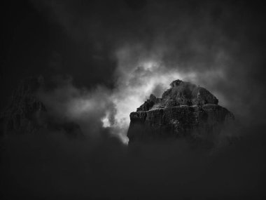 Karanlıktaki kayalık dağın zirvesi güneş ışığıyla aydınlanır siyah beyaz bulutlar arasından, Dolomitler, İtalya