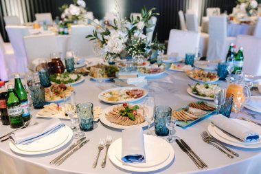 Restoranda beyaz ve mavi çiçeklerle süslenmiş büyük yuvarlak masa. Masada farklı yemekler var. Küçük çocuğun kristali. Lüks dekor.