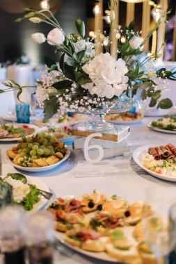 Restoranda beyaz ve mavi çiçeklerle süslenmiş büyük yuvarlak masa. Masada farklı yemekler var. Küçük çocuğun kristali. Lüks dekor.