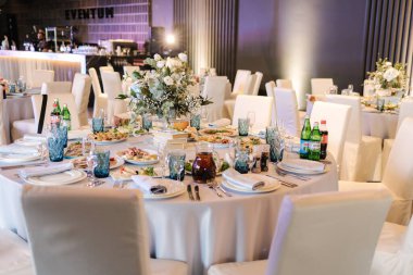 Restoranda beyaz ve mavi çiçeklerle süslenmiş büyük yuvarlak masa. Masada farklı yemekler var. Küçük çocuğun kristali. Lüks dekor.