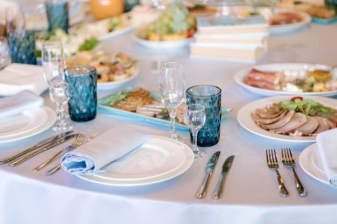 Restoranda beyaz ve mavi çiçeklerle süslenmiş büyük yuvarlak masa. Masada farklı yemekler var. Küçük çocuğun kristali. Lüks dekor.