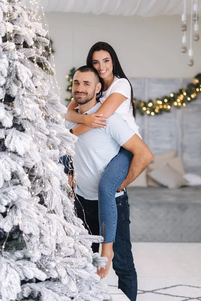 stock image Beautiful woman climbed behind her husbands back and hugged him. Cute couple spend time at home during Christmas. Fit tree.