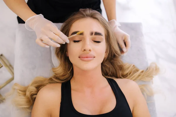 Brow master in protective glowes gives shape to pull out with forceps previously painted with henna eyebrows in a beauty salon. Beautiful blond hair model.