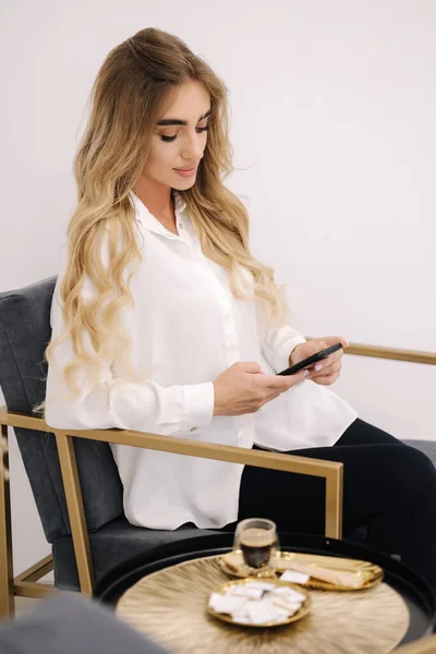化粧品の手順を待っている豪華なモデルは コーヒーを飲み 電話を使用します 美容室の白いインテリア 金髪の女 — ストック写真
