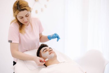 Kozmetik uzmanı spa salonundaki erkeklerin yüzüne kilden maske takıyor..