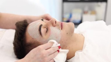 Kozmetik uzmanı, kaplıca salonunda adamın suratındaki ıslak peçeteyle kilden maskeyi yıkar. Güzellik uzmanı, güzellik salonundaki yakışıklı adama dikkat ediyor..