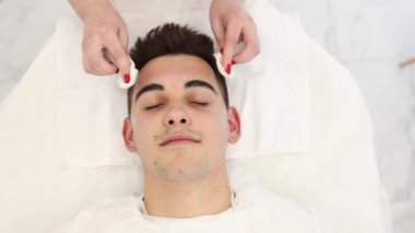 Kozmetik uzmanı, kaplıca salonunda adamın suratındaki ıslak peçeteyle kilden maskeyi yıkar. Güzellik uzmanı, güzellik salonundaki yakışıklı adama dikkat ediyor..