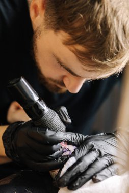 Genç bir profesyonel dövme sanatçısının yakın çekimi, kablosuz dövme makinesinin iğnesiyle deriye siyah mürekkep enjekte ediyor. Elinde dövme var.