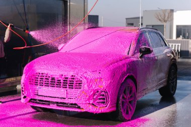 Young woman cleaning her car on self service car wash. Female spread soap using jet sprayer. Pink foam on the car.