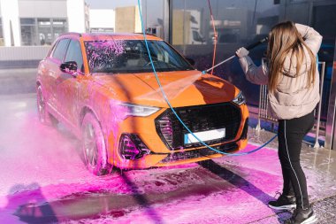 Young woman cleaning her car with a jet sprayer. Self-service car washing. Orange auto.