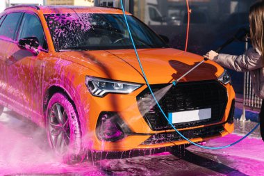 Young woman cleaning her car with a jet sprayer. Self-service car washing. Orange auto.