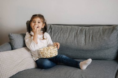 Adorable little girl sits on sofa and watching TV at home. Cute girl eating popcorn. Holiday mood. High quality photo