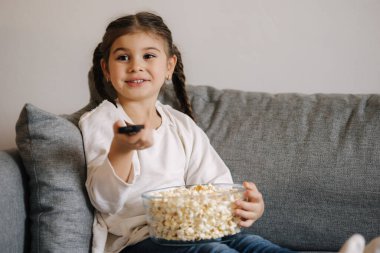 Cute little girl using remote controller of TV and watching cartoons. Four year girl eating popcorn on sofa. High quality photo