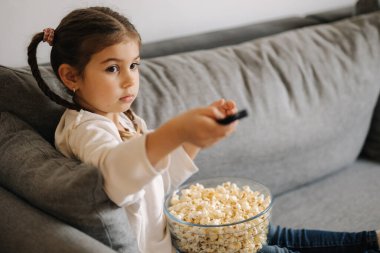 Cute little girl using remote controller of TV and watching cartoons. Four year girl eating popcorn on sofa. High quality photo