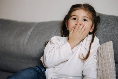 Adorable little girl sits on sofa and watching TV at home. Cute girl eating popcorn. Holiday mood. High quality photo