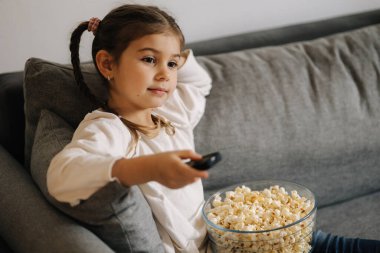 Cute little girl using remote controller of TV and watching cartoons. Four year girl eating popcorn on sofa. High quality photo