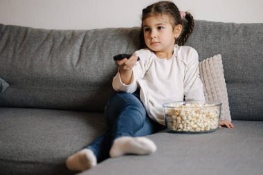 Cute little girl using remote controller of TV and watching cartoons. Four year girl eating popcorn on sofa. High quality photo