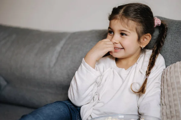 Adorable little girl sits on sofa and watching TV at home. Cute girl eating popcorn. Holiday mood. High quality photo