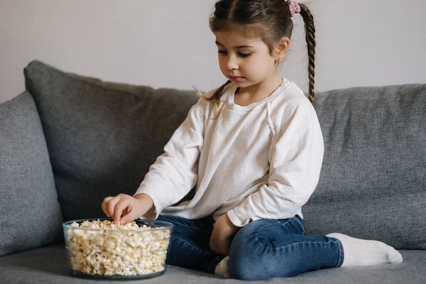 Adorable little girl sits on sofa and watching TV at home. Cute girl eating popcorn. Holiday mood. High quality photo