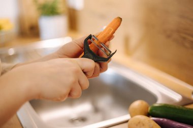 Female peel carrot using vegetable peeler. Close-up of womans hand at kitchen. Preparing food at home. High quality photo