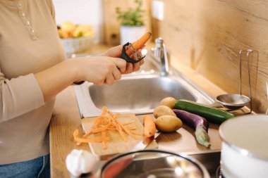 Female peel carrot using vegetable peeler. Close-up of womans hand at kitchen. Preparing food at home. High quality photo