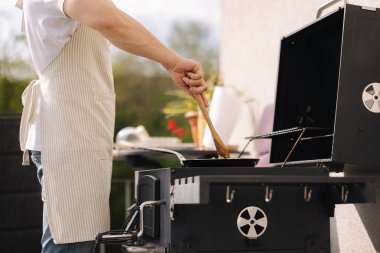 Önlüklü bir adam tavada sebzeleri karıştırmak için tahta kaşık kullanıyor. Izgarada yemek hazırlayın. Yüksek kalite fotoğraf