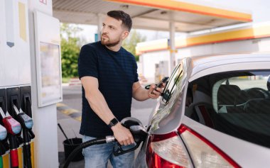 Yakışıklı, sakallı bir adam arabaya yakıt dolduruyor ve benzin istasyonunda dikilirken skorborda bakıyor. Yüksek kalite fotoğraf