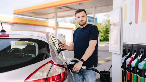 Sakallı adam benzin istasyonunda arabasına yakıt dolduruyor ve akıllı telefonuna bakıyor. İnsanoğlu yakıt fiyatlarını karşılaştırır. Yüksek kalite fotoğraf