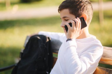 Beyaz tişörtlü, siyah sırt çantalı, parkta bankta oturan ve telefonla konuşan genç bir çocuk..