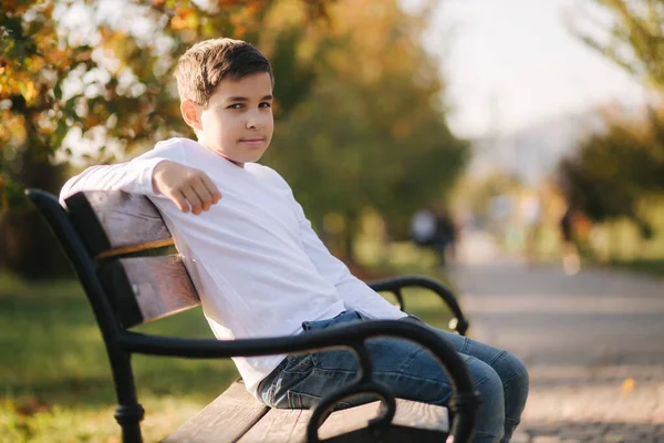 白いセーターを着た10代の少年がベンチに座っている 秋の公園でリュックを持つハンサムな学童 — ストック写真